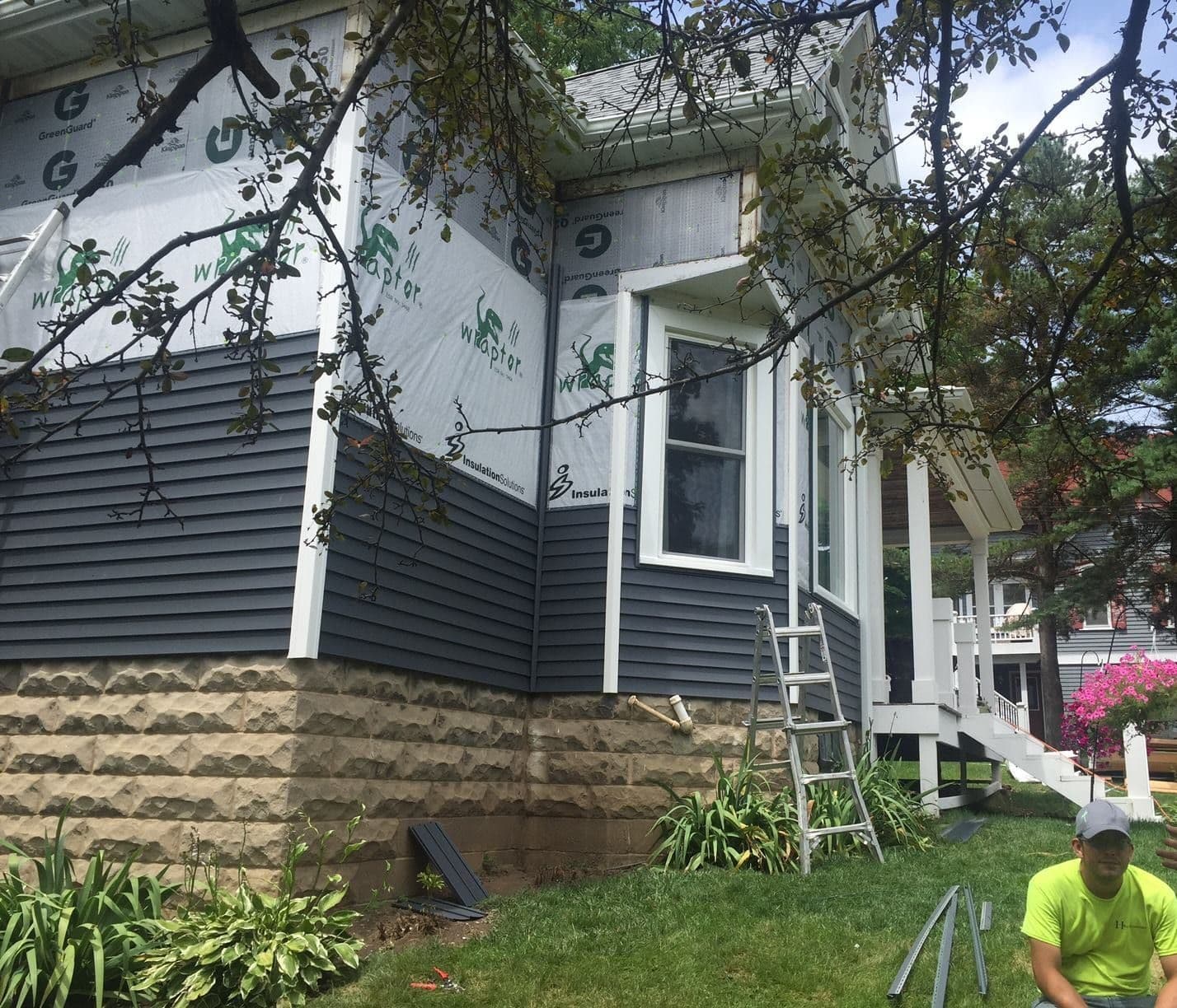 Workers install navy blue vinyl siding on a house with white trim and a tan stone foundation.